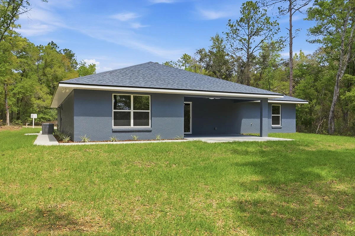 A single-story house painted in blue with a gray shingled roof, situated on a grassy lawn with trees in the background.