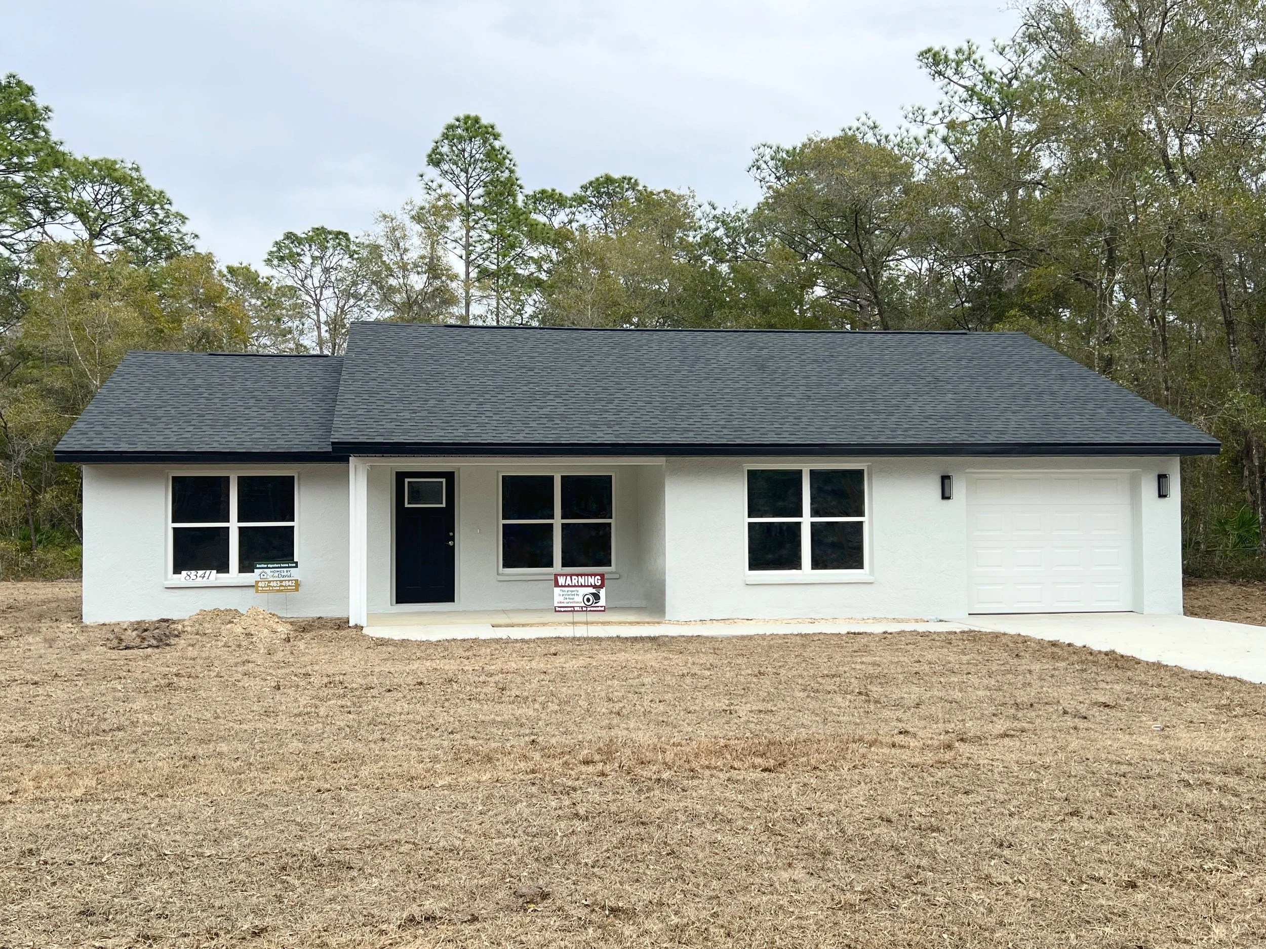 Newly constructed home with white stucco exterior, a black door, black shingles, a single car garage and a covered front porch on a grassy lot with tall trees in the background