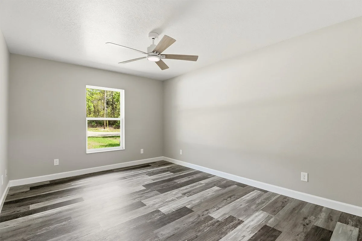 Empty room with light gray walls, a window showing a green outdoor view, wood-look gray flooring, and a ceiling fan.
