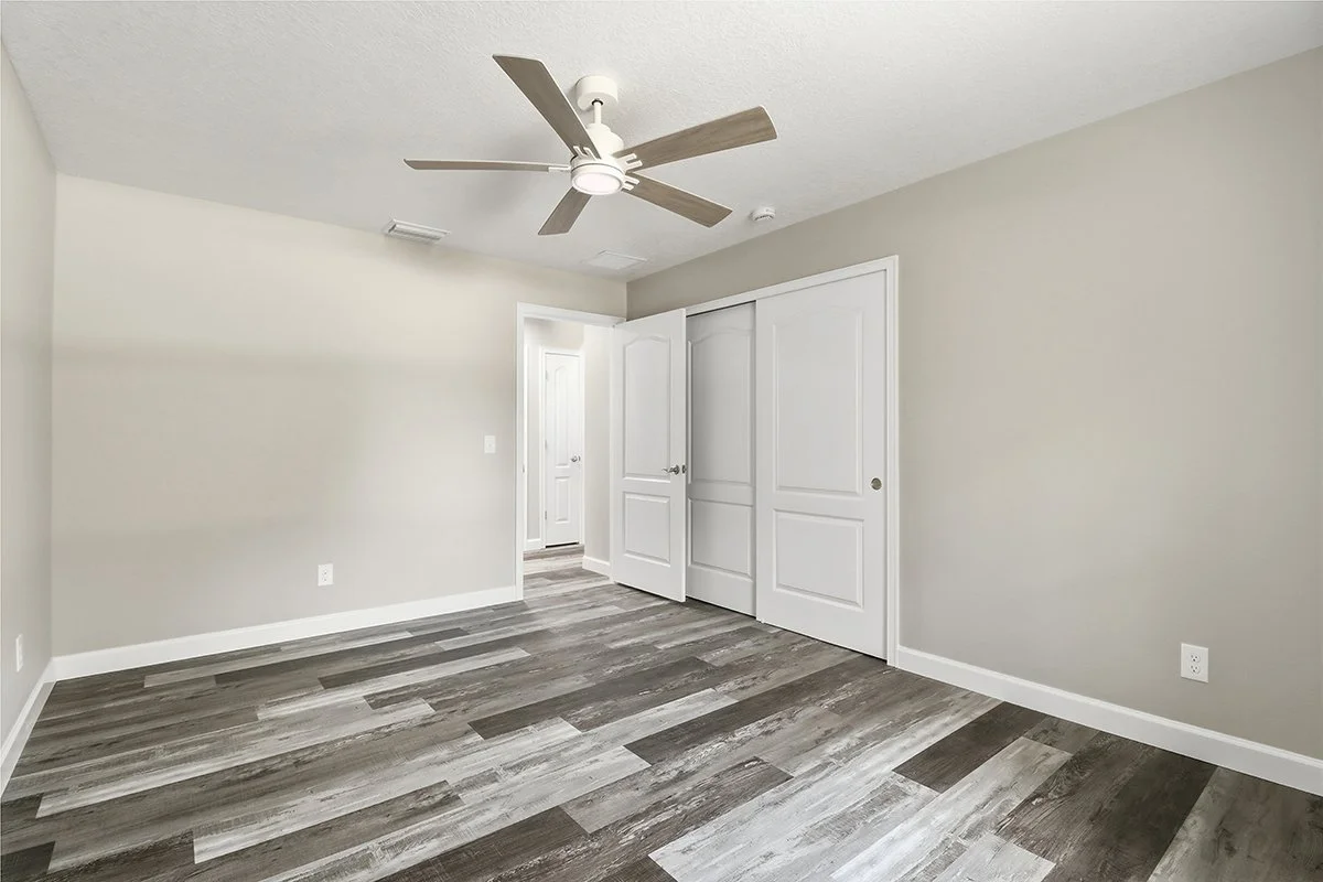 Empty bedroom with light gray walls, gray wood laminate flooring, white ceiling with a modern ceiling fan, and a closet with white sliding doors.