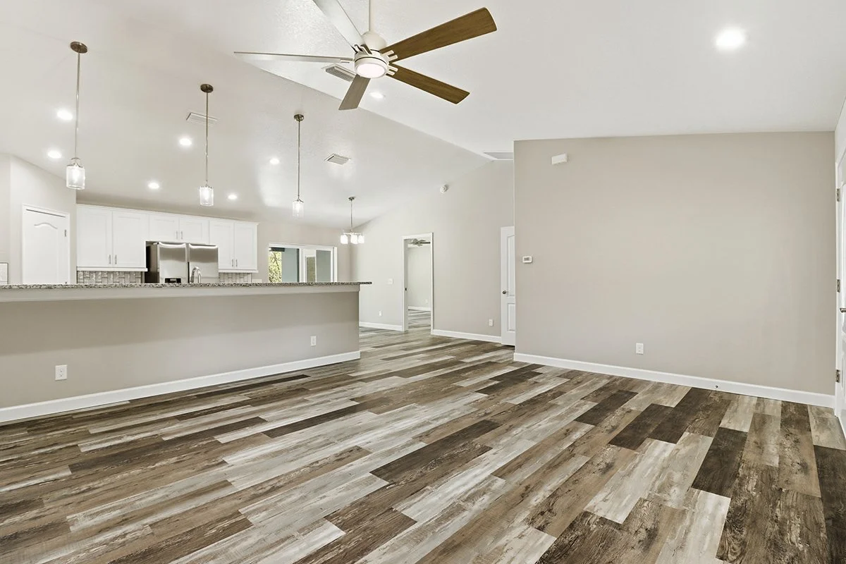 Empty living room with wood-look flooring, beige walls, white baseboards, and a ceiling fan with wooden blades. The background shows an open kitchen with white cabinets, pendant lighting, and a stainless steel refrigerator.