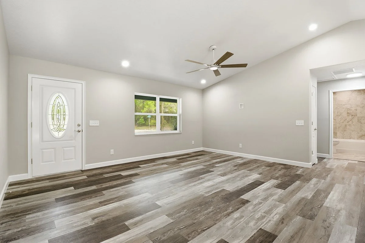 Empty living room with gray walls, wood laminate flooring, ceiling fan, window, and visible bathroom in background.