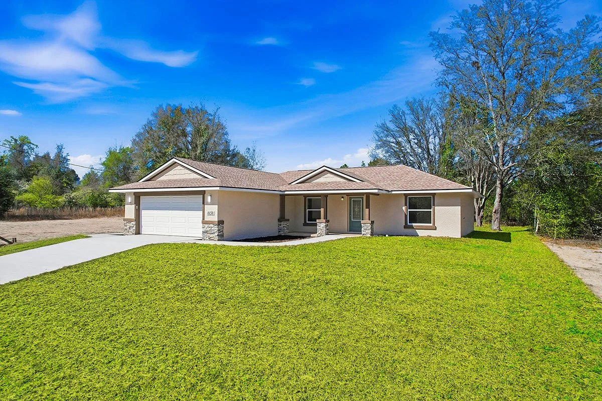 A single-story modern house with a two-car garage, beige exterior, stone accents on the columns, front porch, and green front yard under a bright blue sky.