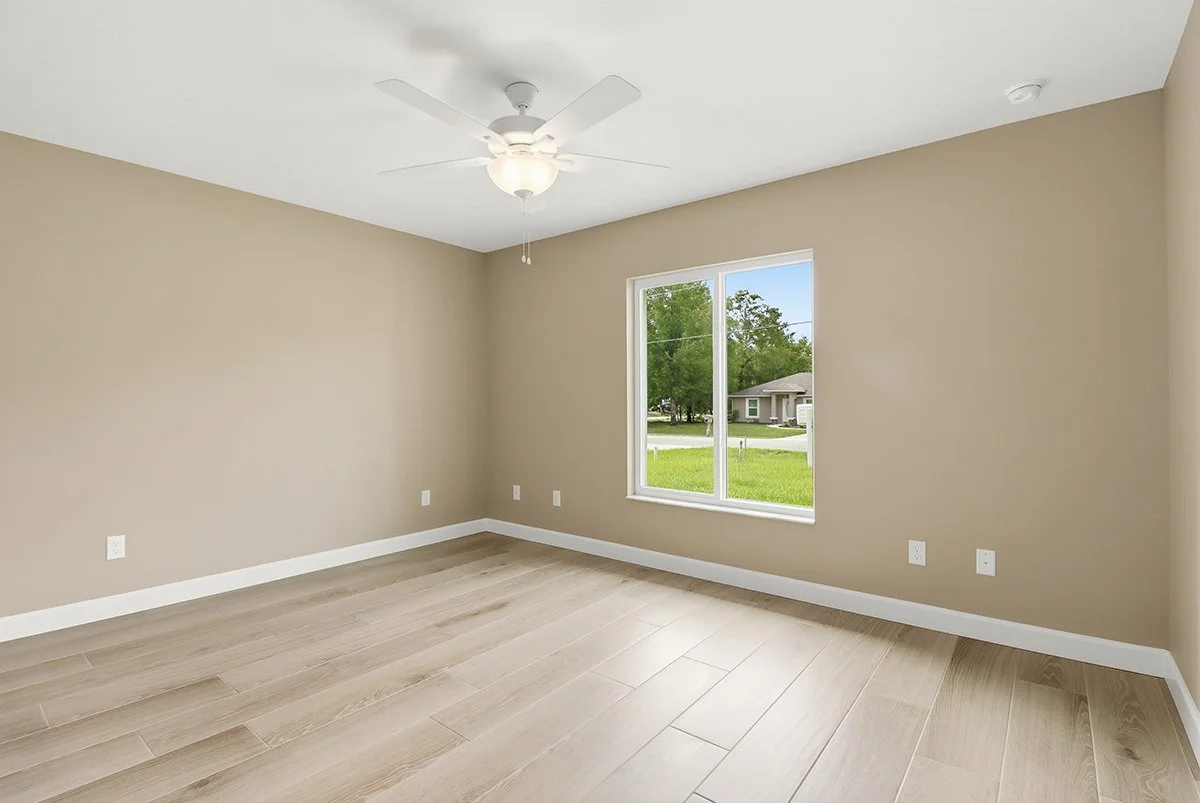 Empty beige room with a large window, ceiling fan, and light-colored wooden flooring.