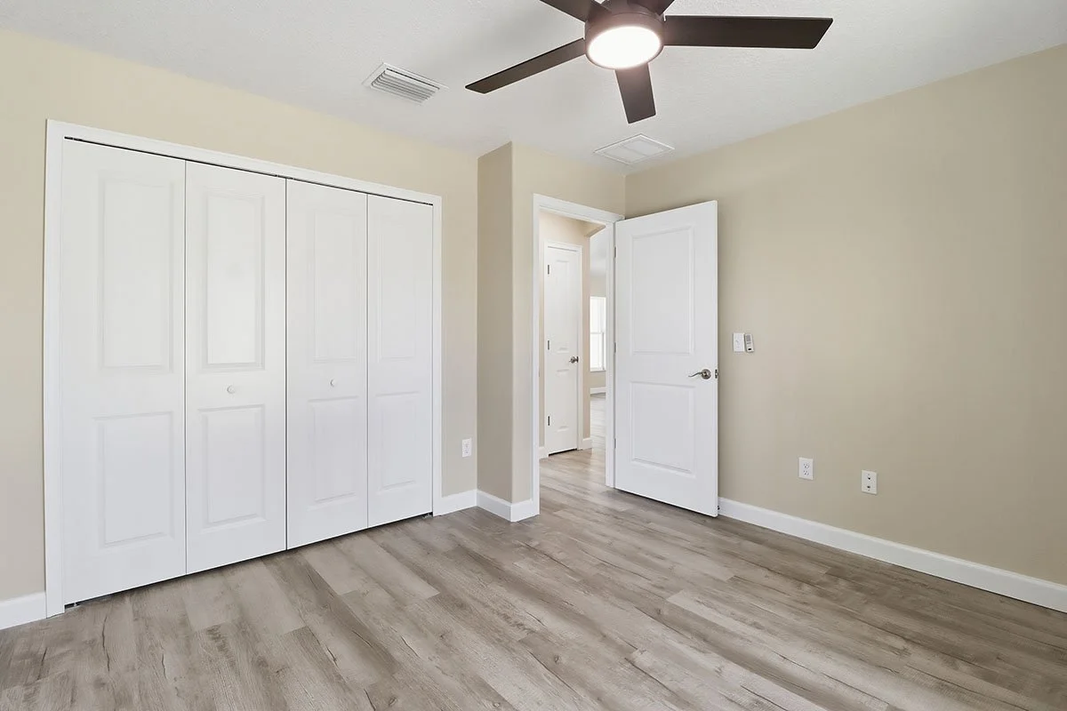 Empty bedroom with beige walls, white closet doors, a ceiling fan, hardwood floor, and open door leading to another room.
