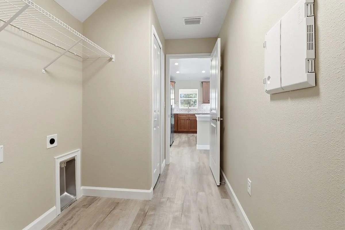 Empty laundry room with beige walls, white wire shelf, pet door, electrical outlets, and a view into a kitchen with wooden cabinets and a window.
