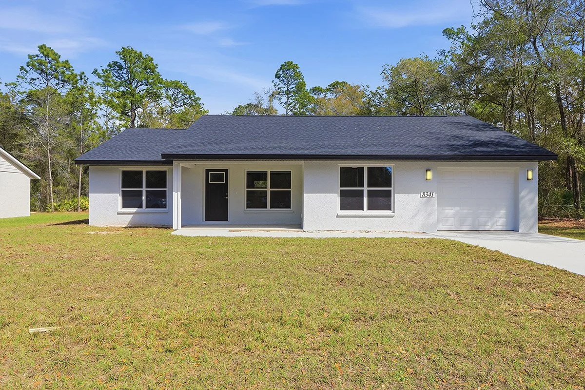 Single story white house in Crystal River, Florida with a black roof and black door that has a single car garage with a white garage door. The front of the house has three sets of 4-pane windows. Tall trees and blue sky are in the background