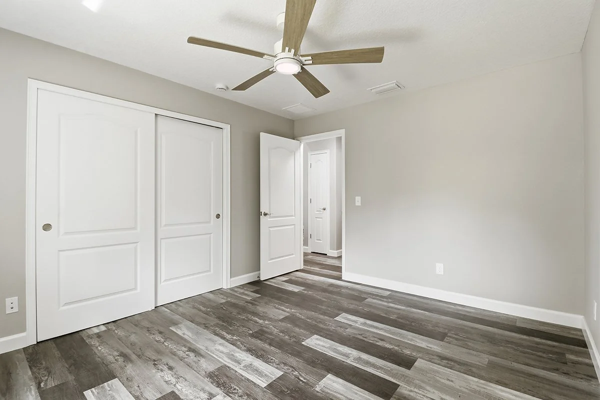 Empty bedroom with gray walls, wood-look flooring, white baseboards, ceiling fan, and closet doors.