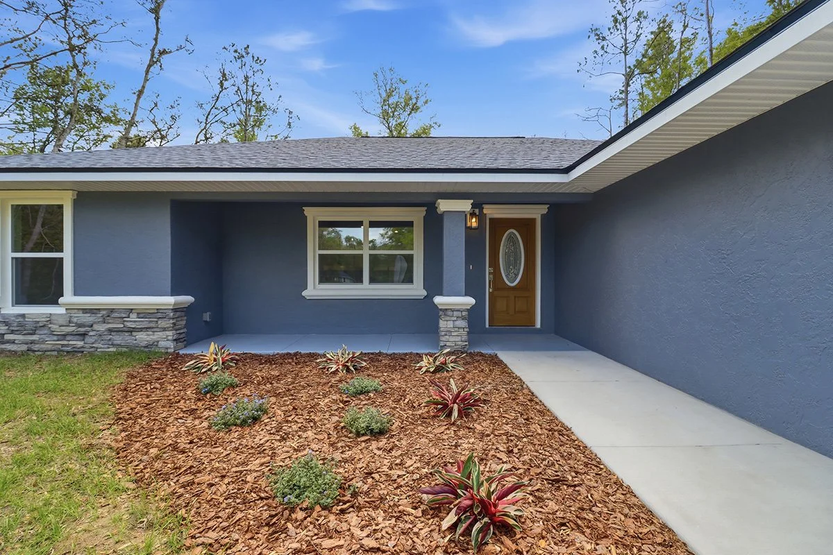 Front view of a modern house with a blue exterior, stone accents, a brown front door, and a small garden with plants and mulch.
