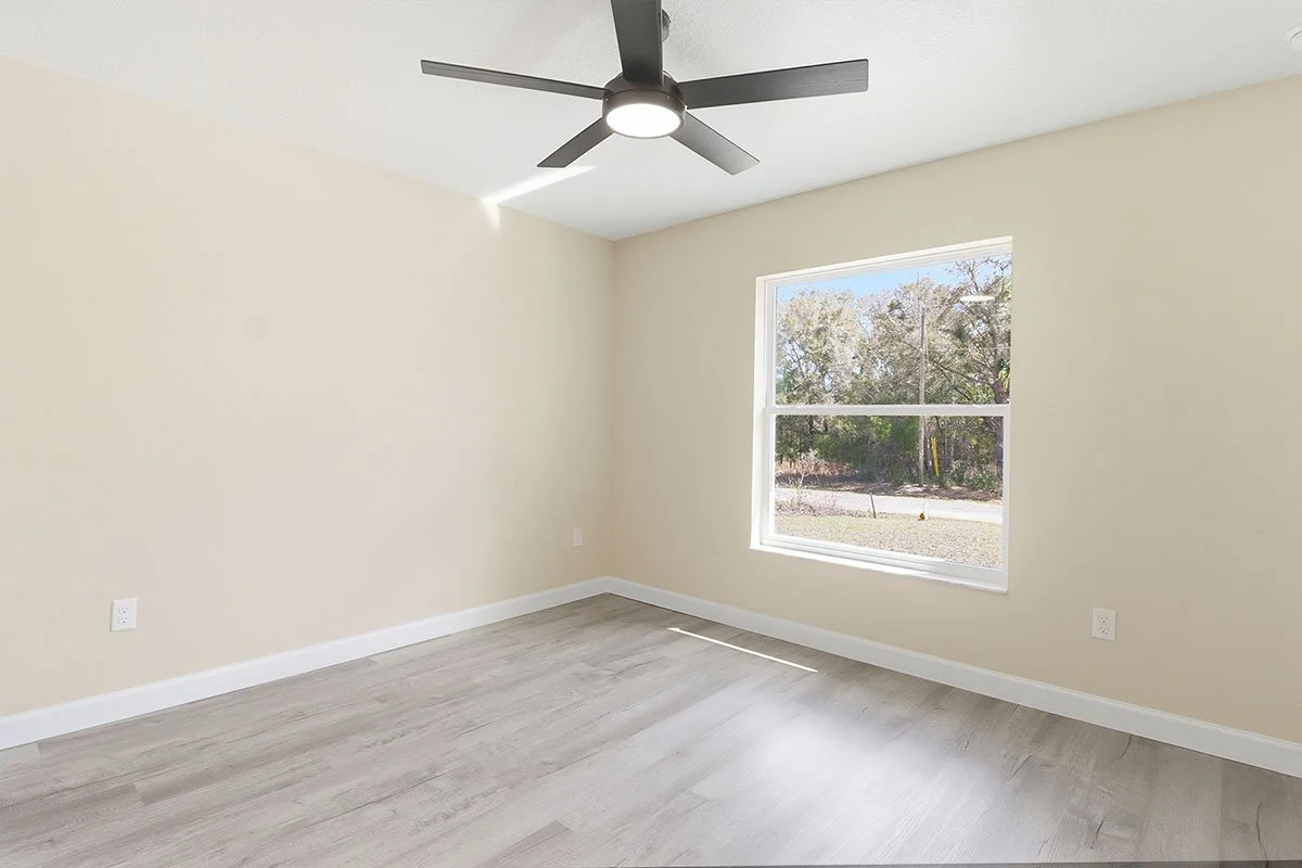Empty room with beige walls, a single window, light wood flooring, white baseboards, and a ceiling fan with lights.