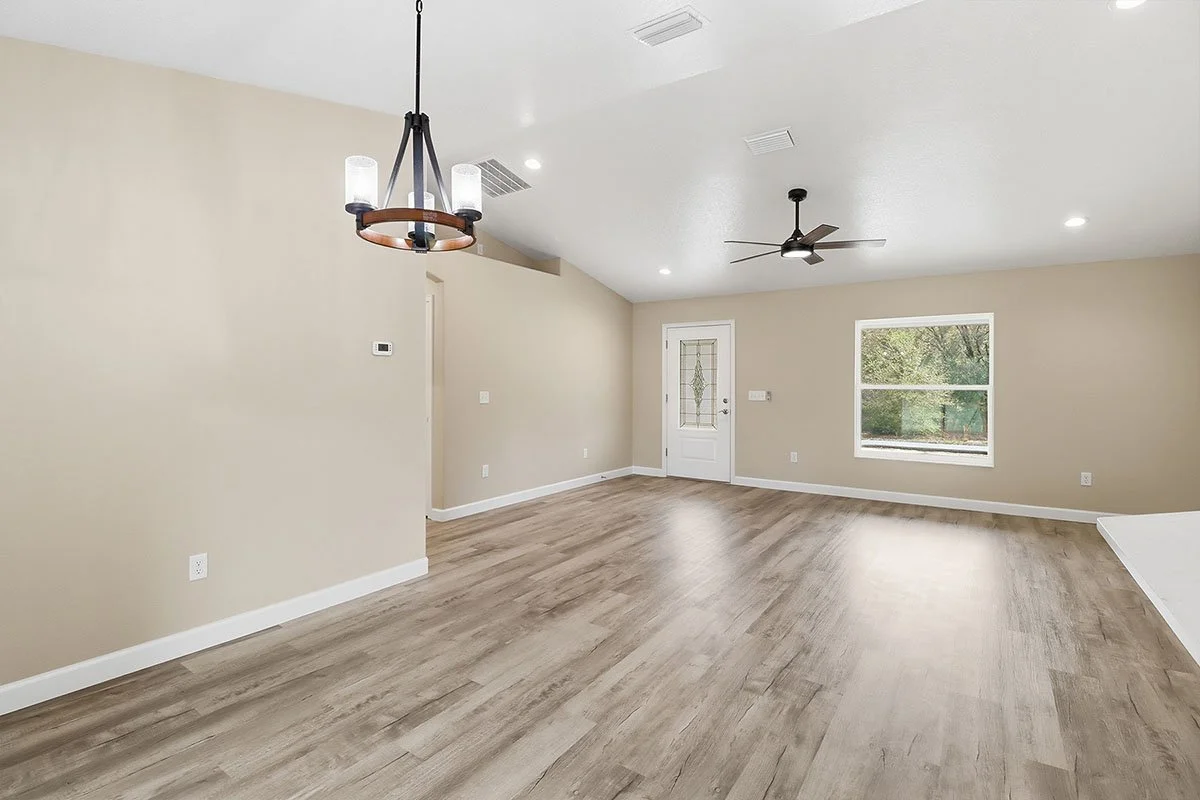 Empty living room with beige walls, wood flooring, a ceiling fan, a chandelier, a window, and a door.