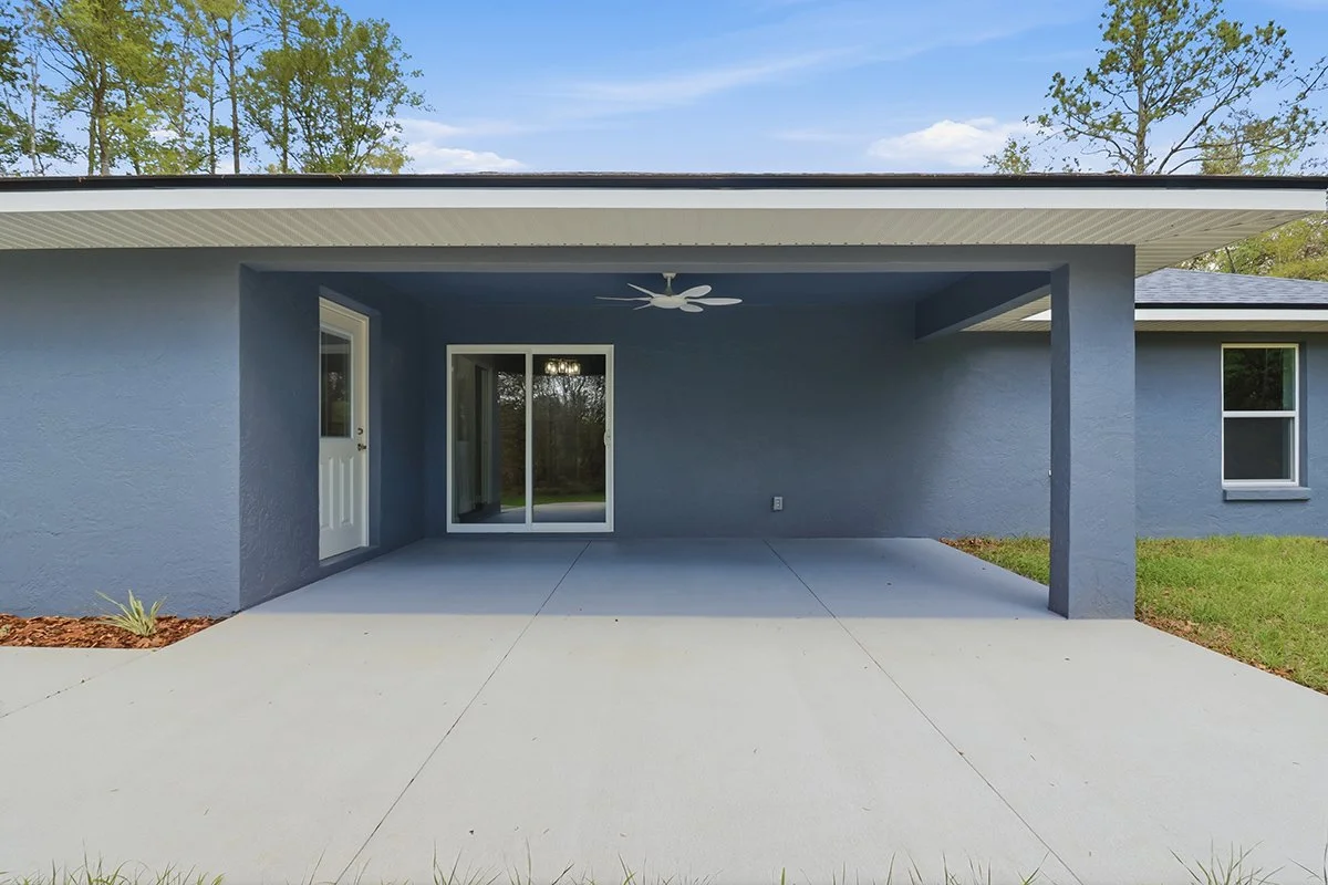 Backyard patio with concrete flooring, dark blue painted walls, a white door, a sliding glass door, and a ceiling fan.