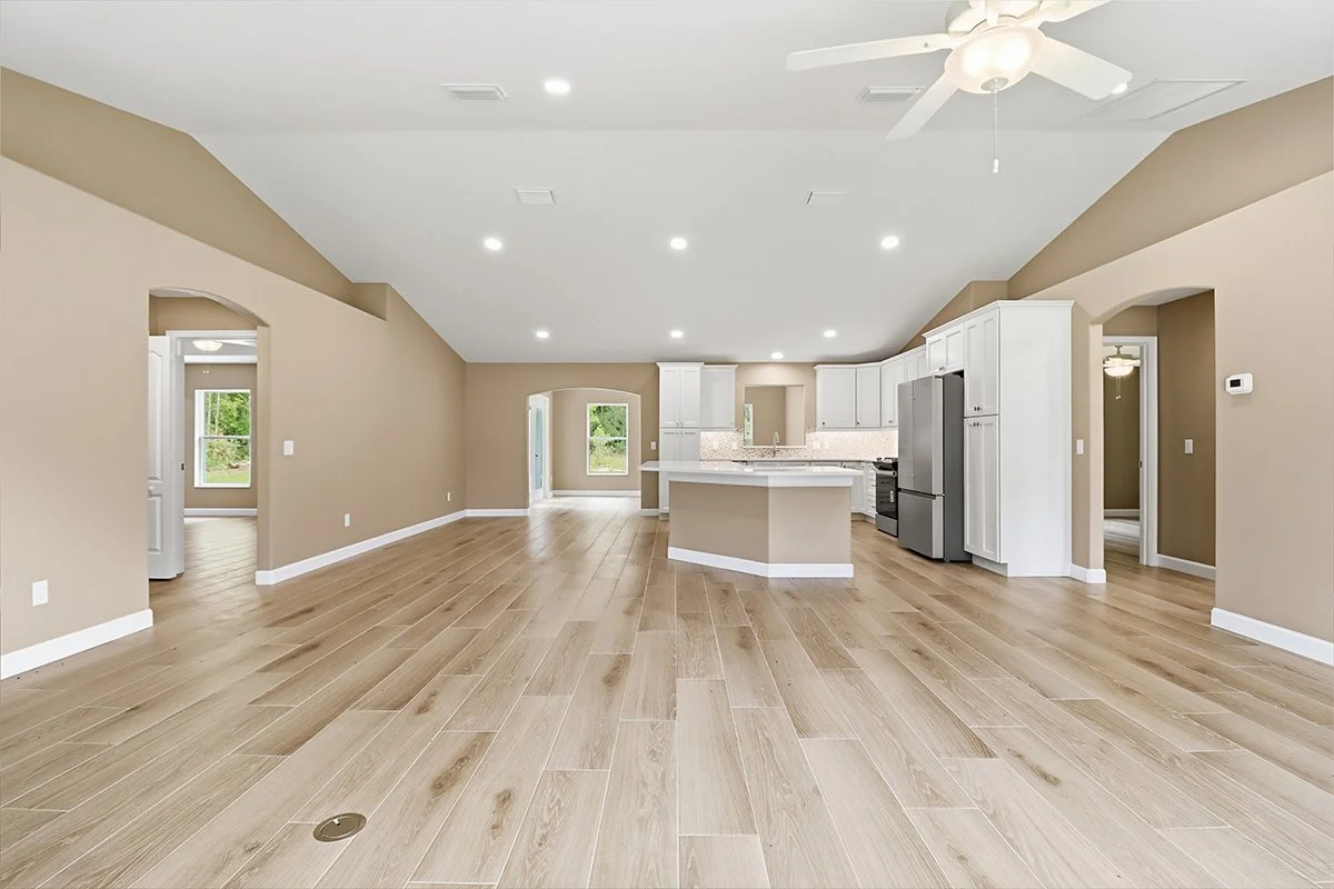 Open concept living area with a kitchen featuring white cabinets, stainless steel appliances, a kitchen island, and light wood flooring. The room has beige walls, a ceiling fan, and ample natural light.
