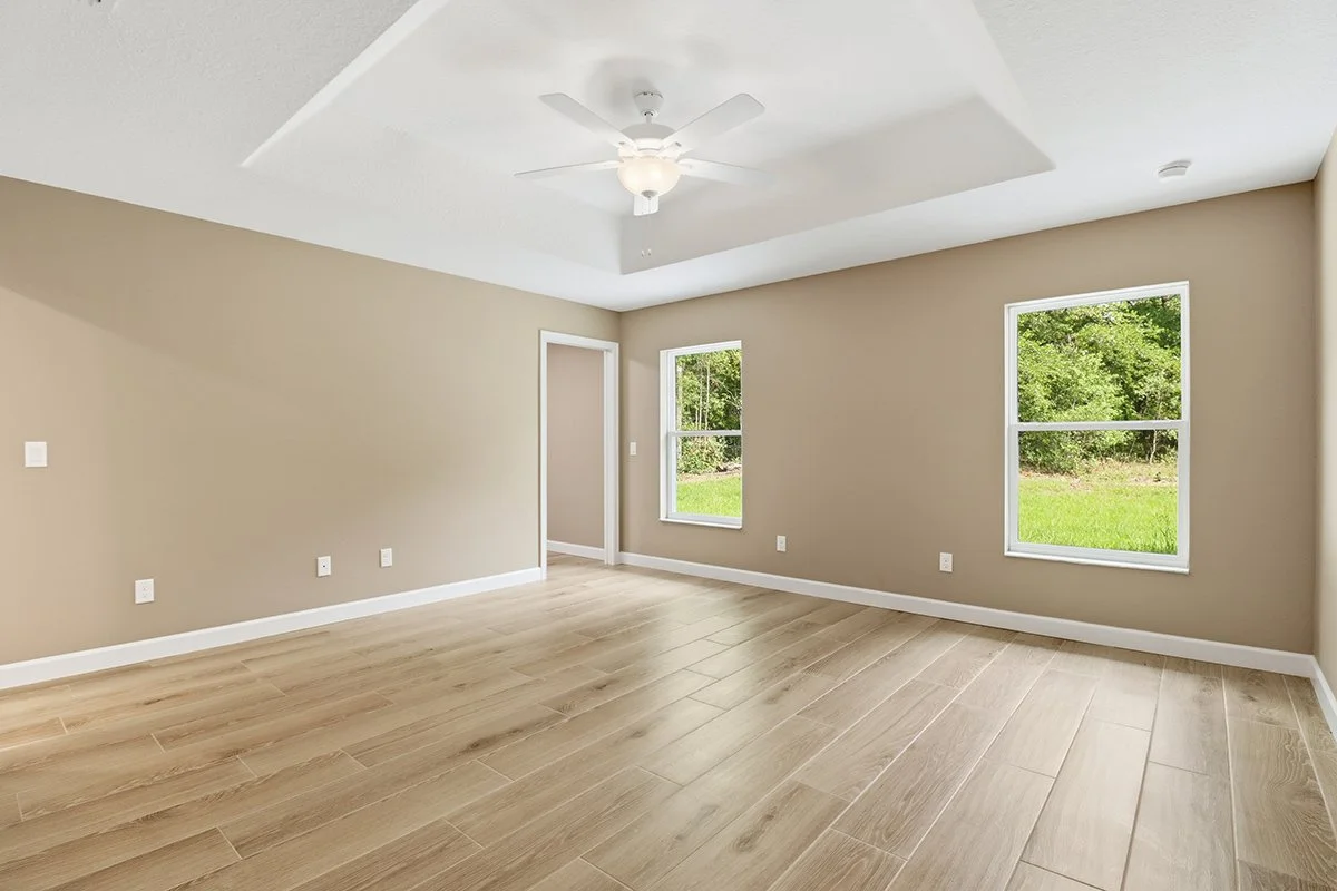 Empty room with beige walls, light wood flooring, two large windows showing greenery outside, a white ceiling fan, and a doorway.