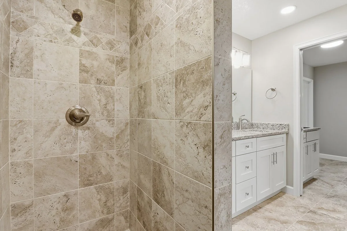A bathroom with a walk-in shower covered in beige stone tiles, a white vanity with a granite countertop, a mirror, and wall-mounted lights.