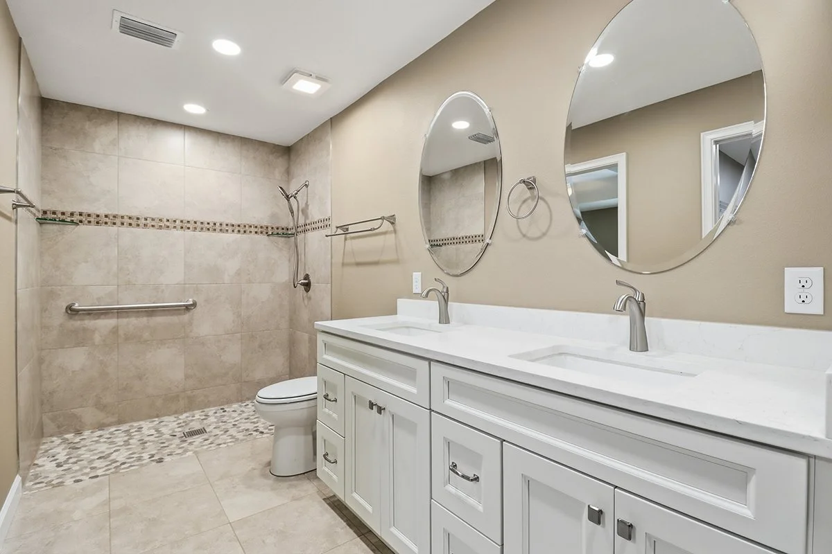 Bathroom with double vanity sinks, large round mirrors, beige walls, beige tiled shower with grab bar and small mosaic accents, and a toilet. Recessed ceiling lights and a towel ring are visible.
