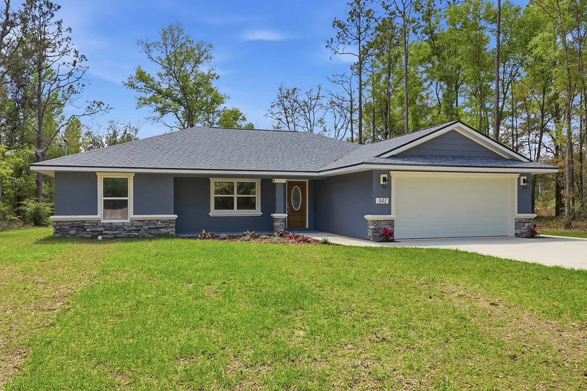 Single story ranch style home in Citrus County Florida painted dark blue with white trim, stone accents, blue roof, and two car garage with white door. Grassy front yard and concrete driveway. Tall trees and blue sky in the background.