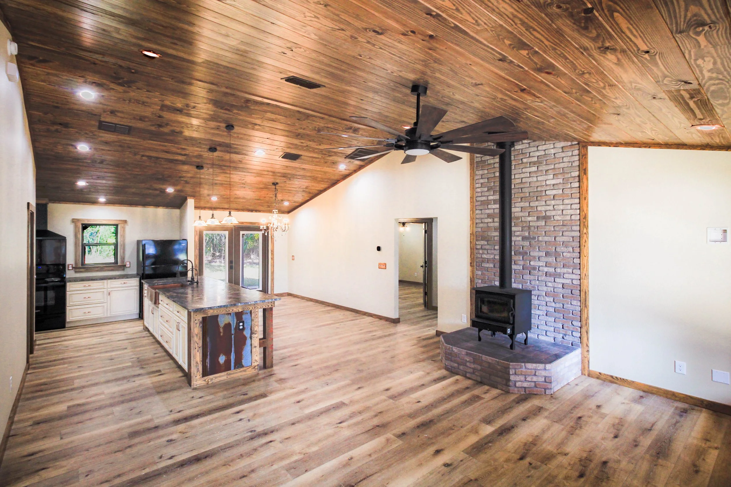 Interior of single story open floor plan cabin with tongue and groove ceiling, hardwood floors, large kitchen island, and fireplace