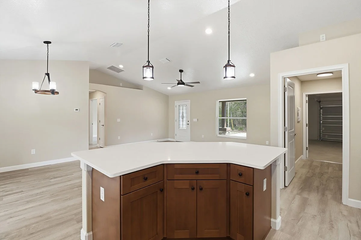 Modern kitchen with a white countertop island, wooden cabinets, hanging pendant lights, a ceiling fan, and a window overlooking an outdoor area.