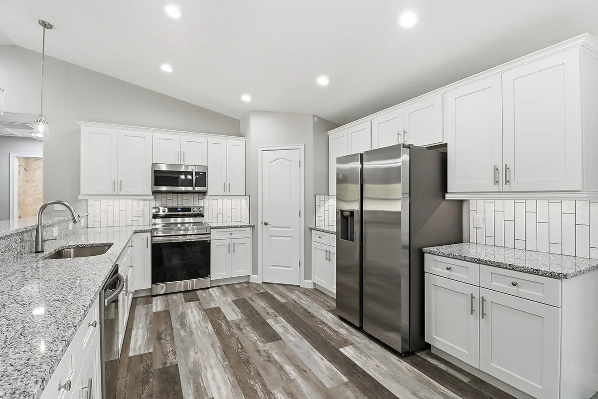Modern kitchen with white cabinets, stainless steel appliances, granite countertops, wood-look flooring, and white tiled backsplash.