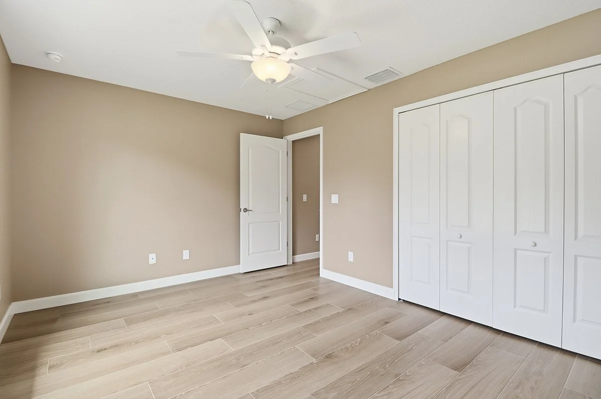 Empty bedroom with beige walls, wood-like floor, white ceiling fan, open closet doors, and a door leading to another room.