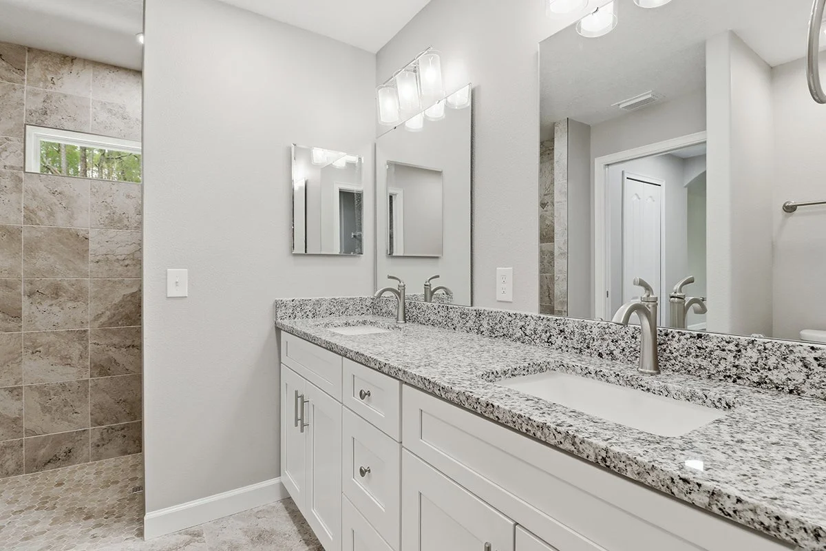 Bathroom with a dual sink granite countertop, white cabinetry, large mirror, and beige tile shower area.