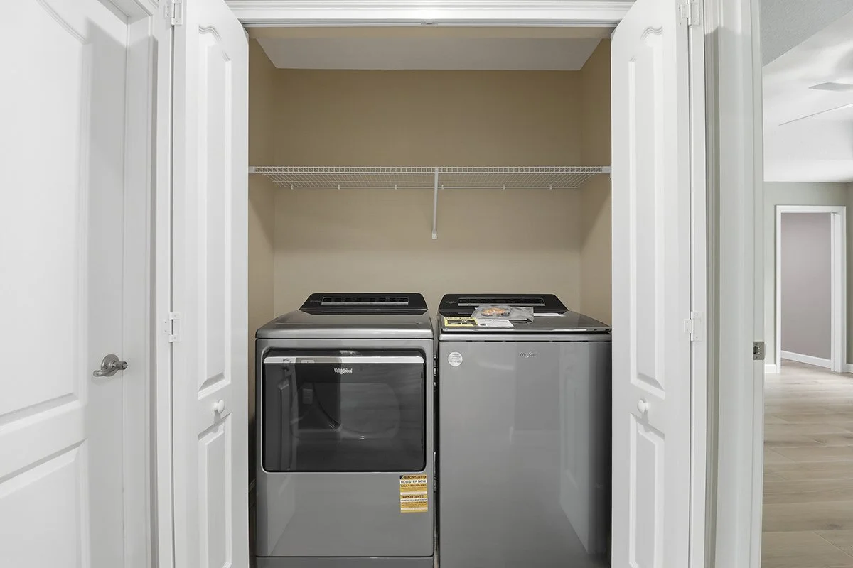 A laundry closet with a washing machine on the left and a dryer on the right, enclosed by white double doors, with a wire shelf above them.