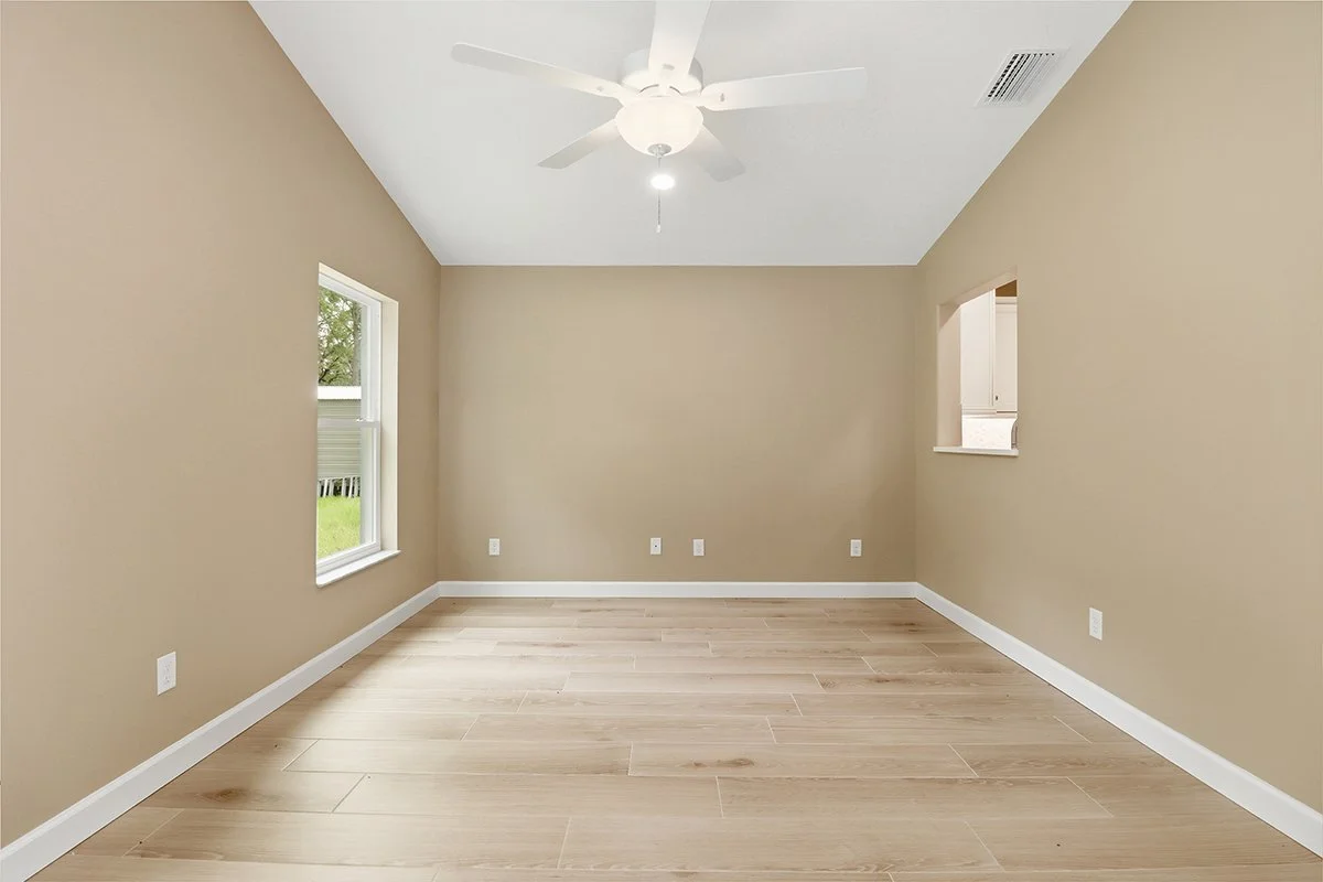 Empty room with beige walls, light wood flooring, two windows, ceiling fan, and a small open window to an adjacent room.