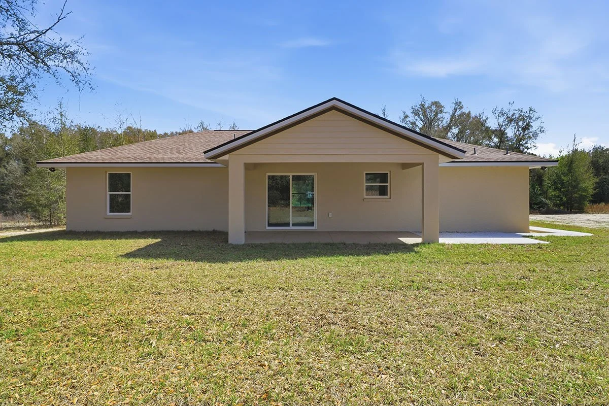 Rear view of a new single-story house with beige siding, a brown shingle roof, and a covered patio area with two support columns. The backyard has a grassy lawn and some trees in the background.