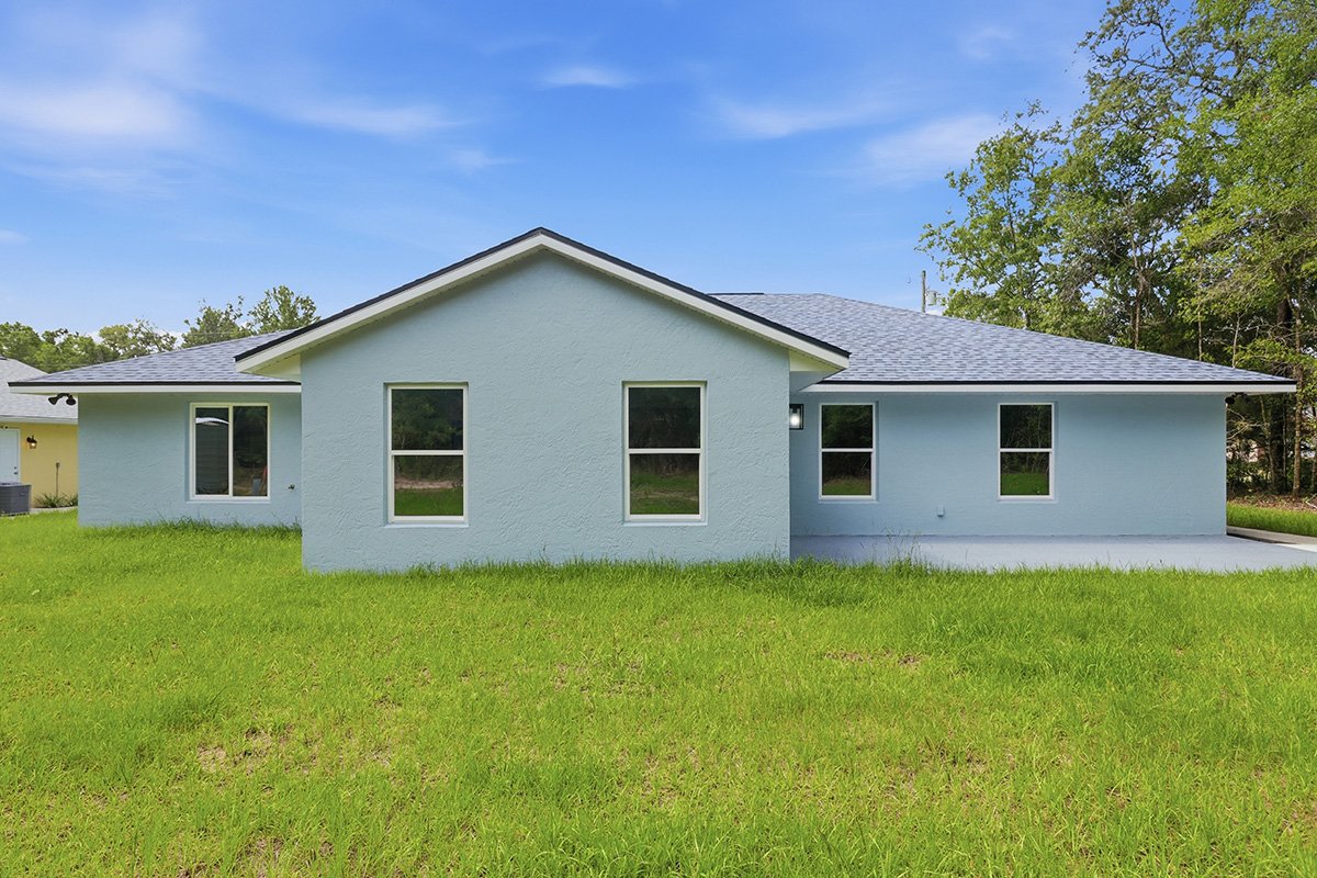 A light blue, single-story house with multiple windows, a gray shingle roof, and a grassy yard under a partly cloudy sky.