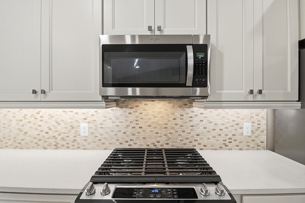 Kitchen with a microwave above a gas stove, beige tile backsplash, white cabinetry, and electrical outlets.