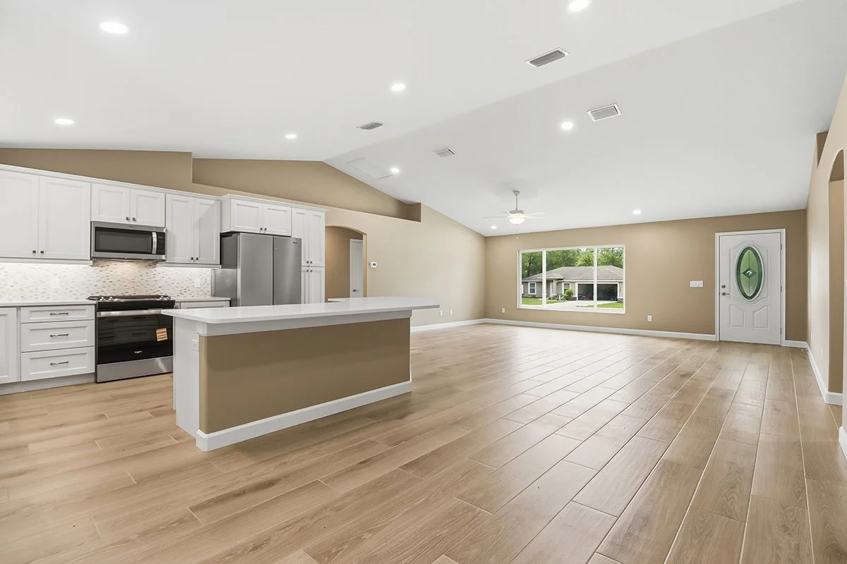 Empty living room and kitchen with light-colored wooden floors, beige walls, large window, white ceiling with recessed lighting, and a white front door with a green oval window.