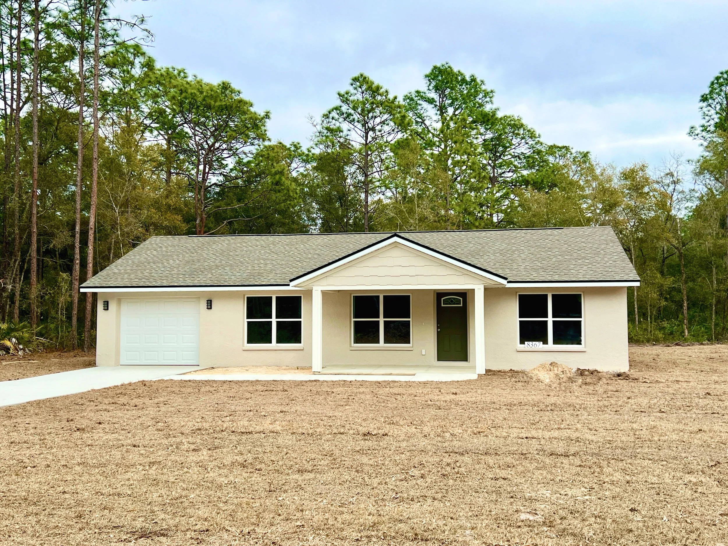 Single story newly constructed with a single car garage, a small covered front porch and a concrete driveway. The home is painted a creamy beige color with a dark green front door and being shingled roof and sits on a grassy lot.