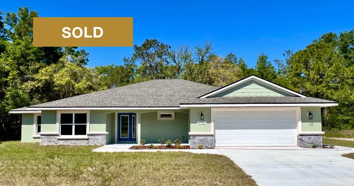 Newly constructed home with sage green stucco exterior, gray stone accents, covered front porch and two car garage