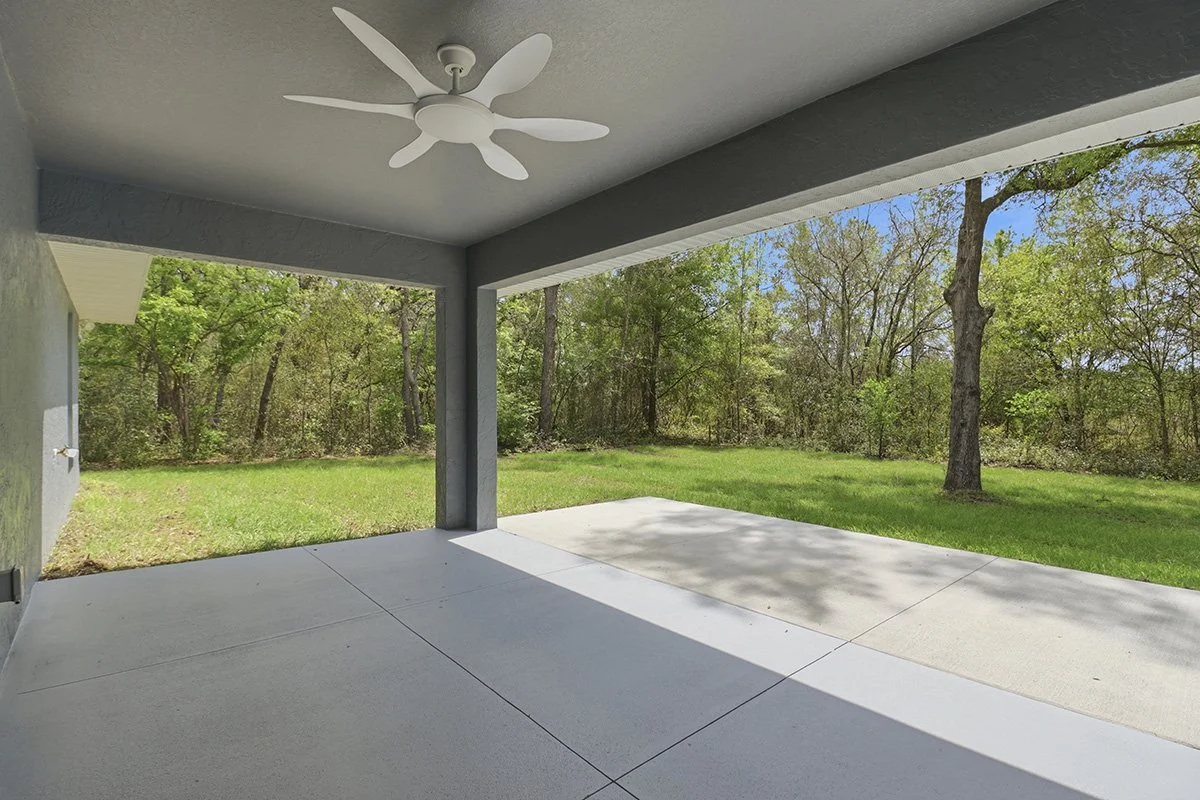 Covered patio with a white ceiling fan, concrete floor, gray painted walls, and a backyard with trees and grass.