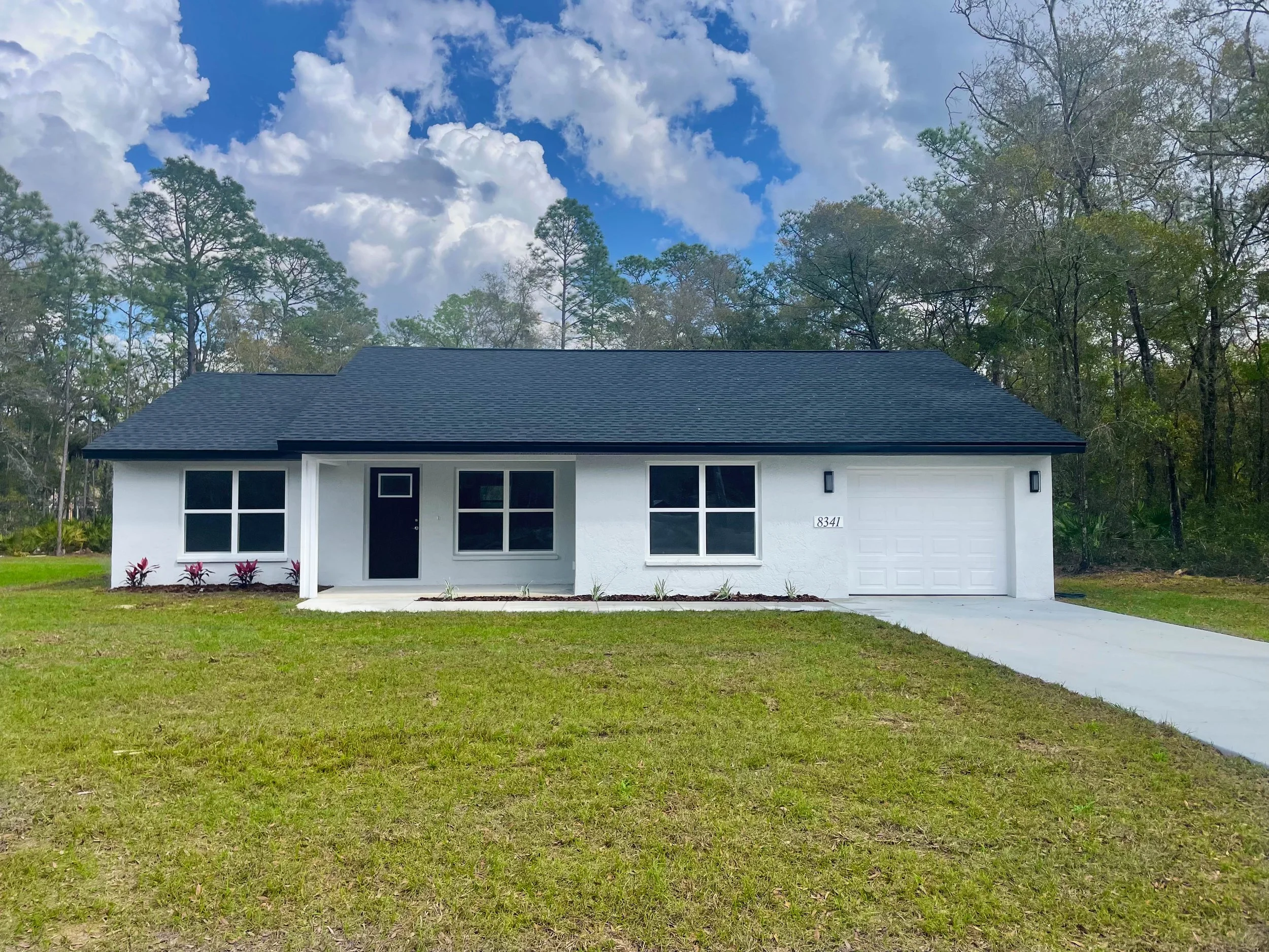 Newly constructed home with white stucco exterior, a black door, black shingles, a single car garage and a covered front porch on a grassy lot with tall trees in the background