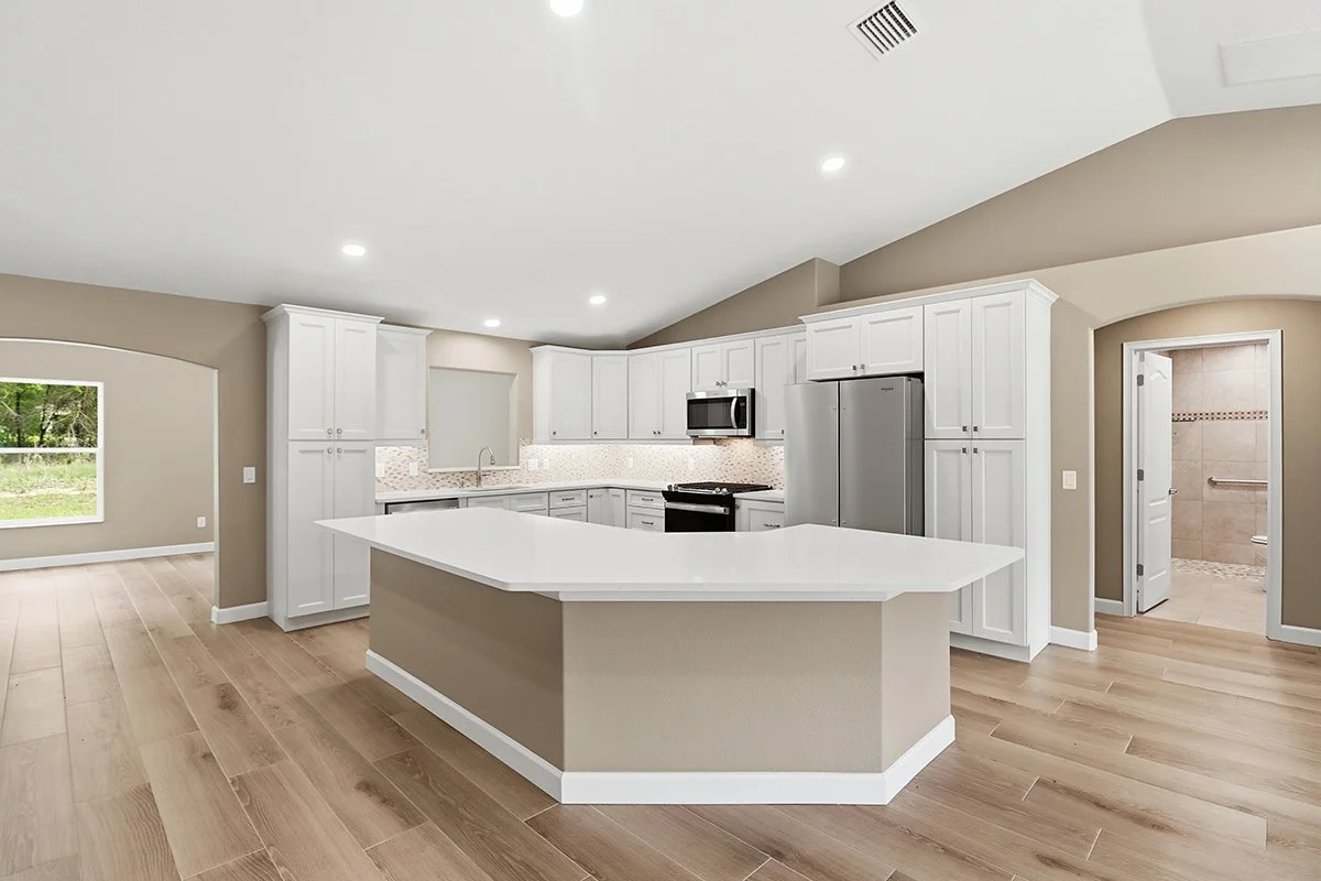 Modern kitchen with white cabinets, stainless steel refrigerator, microwave, black stove, and a large white island with a beige base. Light wood flooring and beige walls, with a view of a bathroom through an open door.