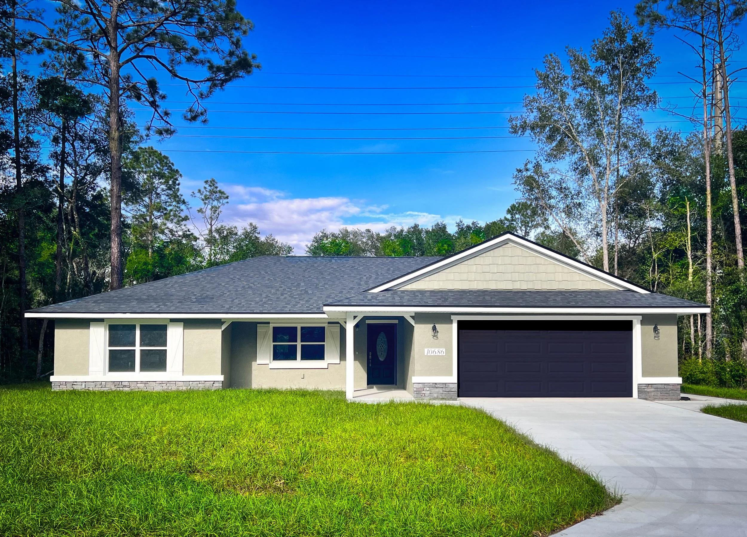 Single-story stucco house with pale greenish gray paint, a black garage door, and a black front door. The roof has dark shingles, stone accents and white trim, with a two-car garage and sits on a grassy lot with concrete driveway