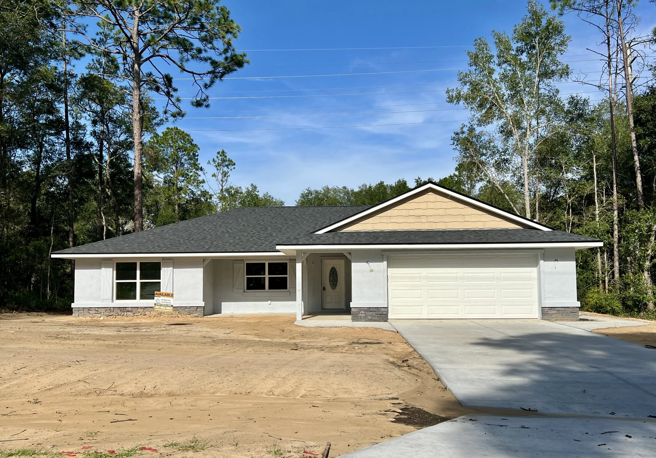 Single-story stucco house under construction with white primer exterior, featuring dark roof shingles, stone accents along the base, two-car garage with white door, and an unfinished dirt yard.