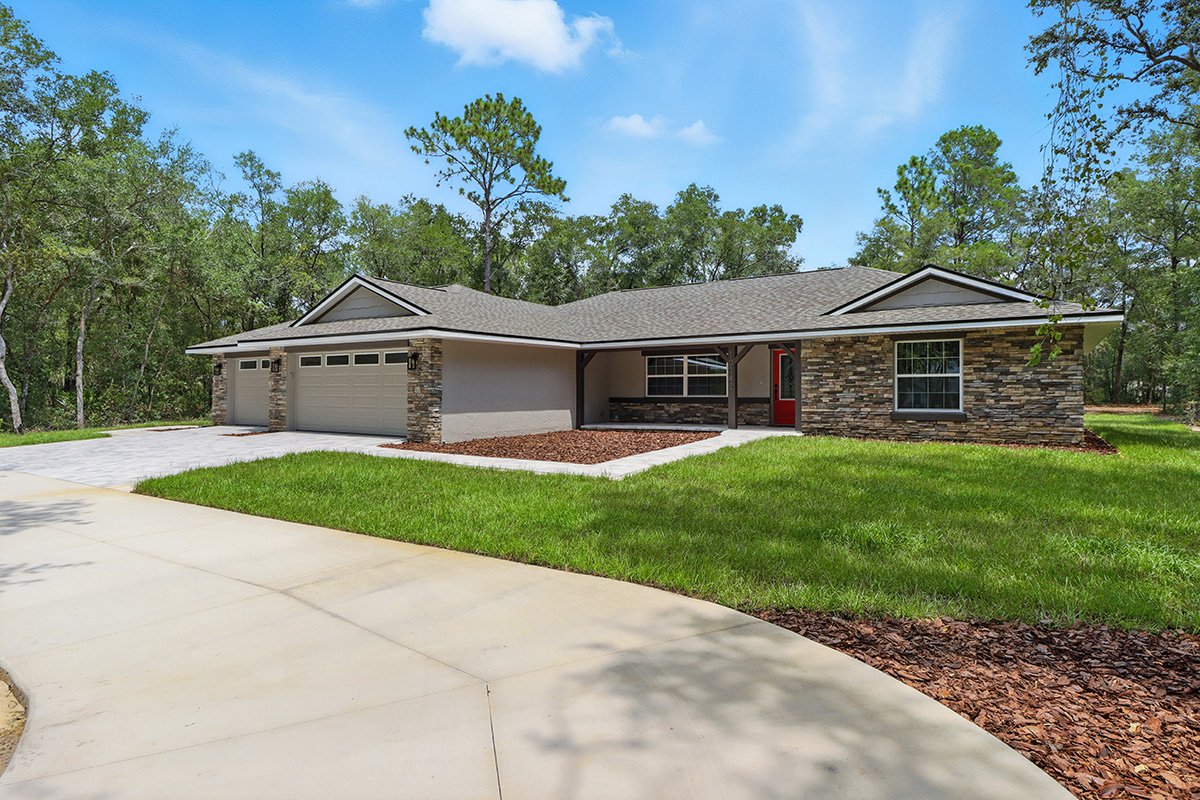 Single-story house with a gray roof, brick and light gray exterior walls, red front door, attached three-car garage, and a front yard with green grass and mulch, surrounded by trees under a blue sky.
