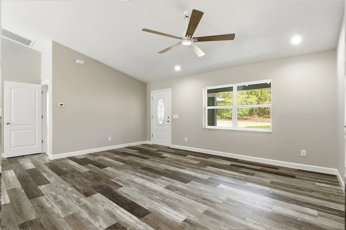 Empty living room with light gray walls, wood-look flooring, a white front door with window, a large window showing greenery outside, a ceiling fan with wooden blades, and a ceiling light.