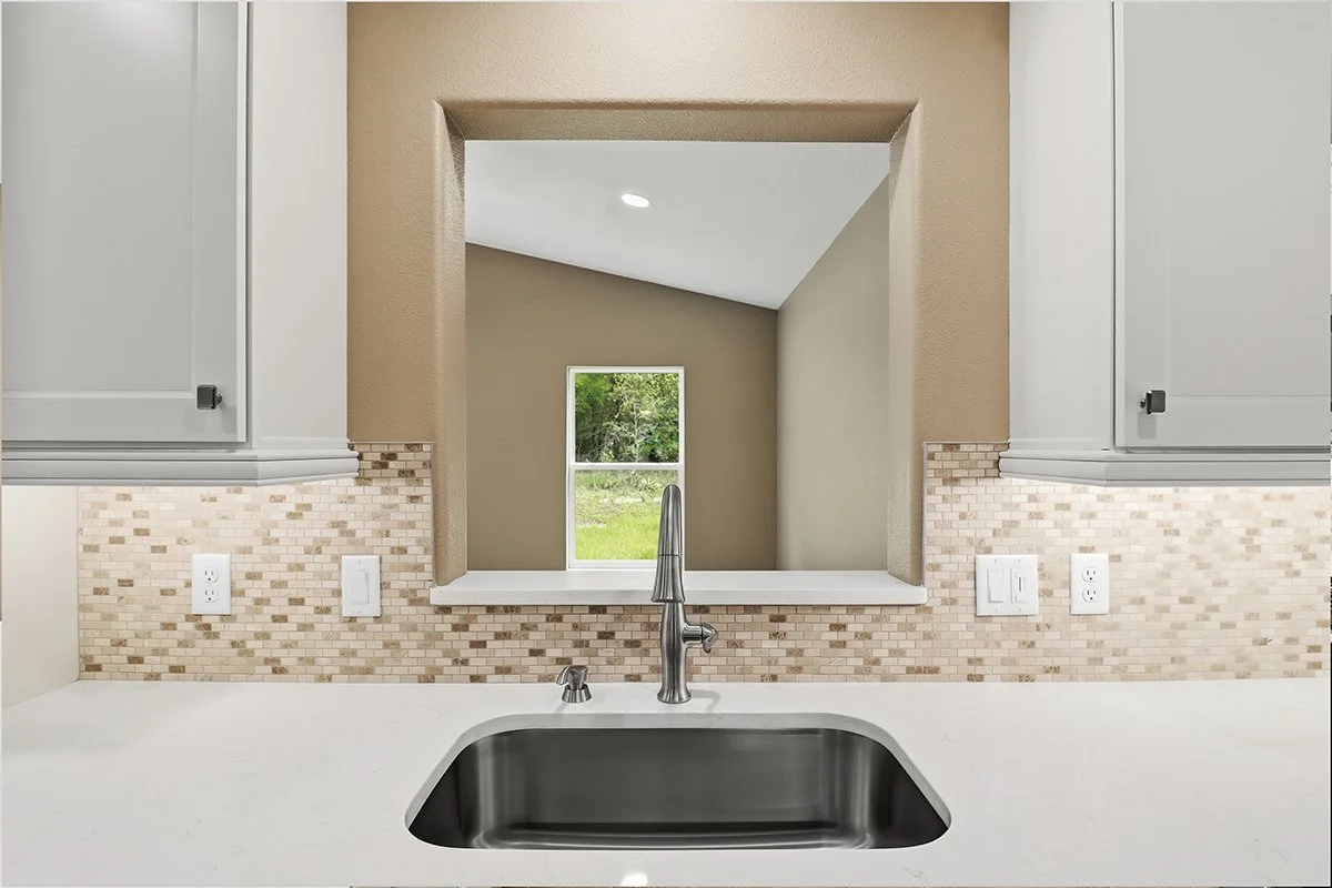 Kitchen sink with stainless steel faucet, beige mosaic tile backsplash, and white countertop, view through window opening to a room with beige wall and window showing green outdoors.
