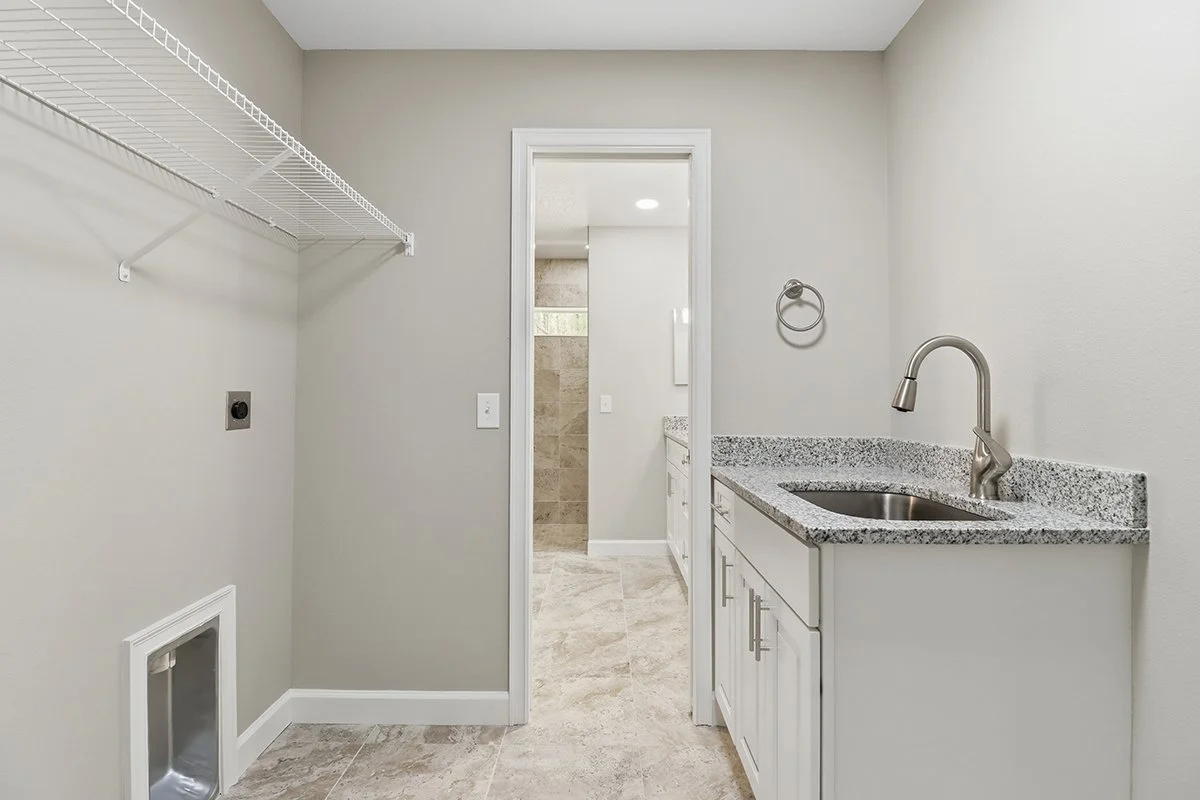 Empty laundry room with a wire shelf, a small pet door, a sink with a granite countertop, and a doorway leading to a bathroom with a tiled shower.