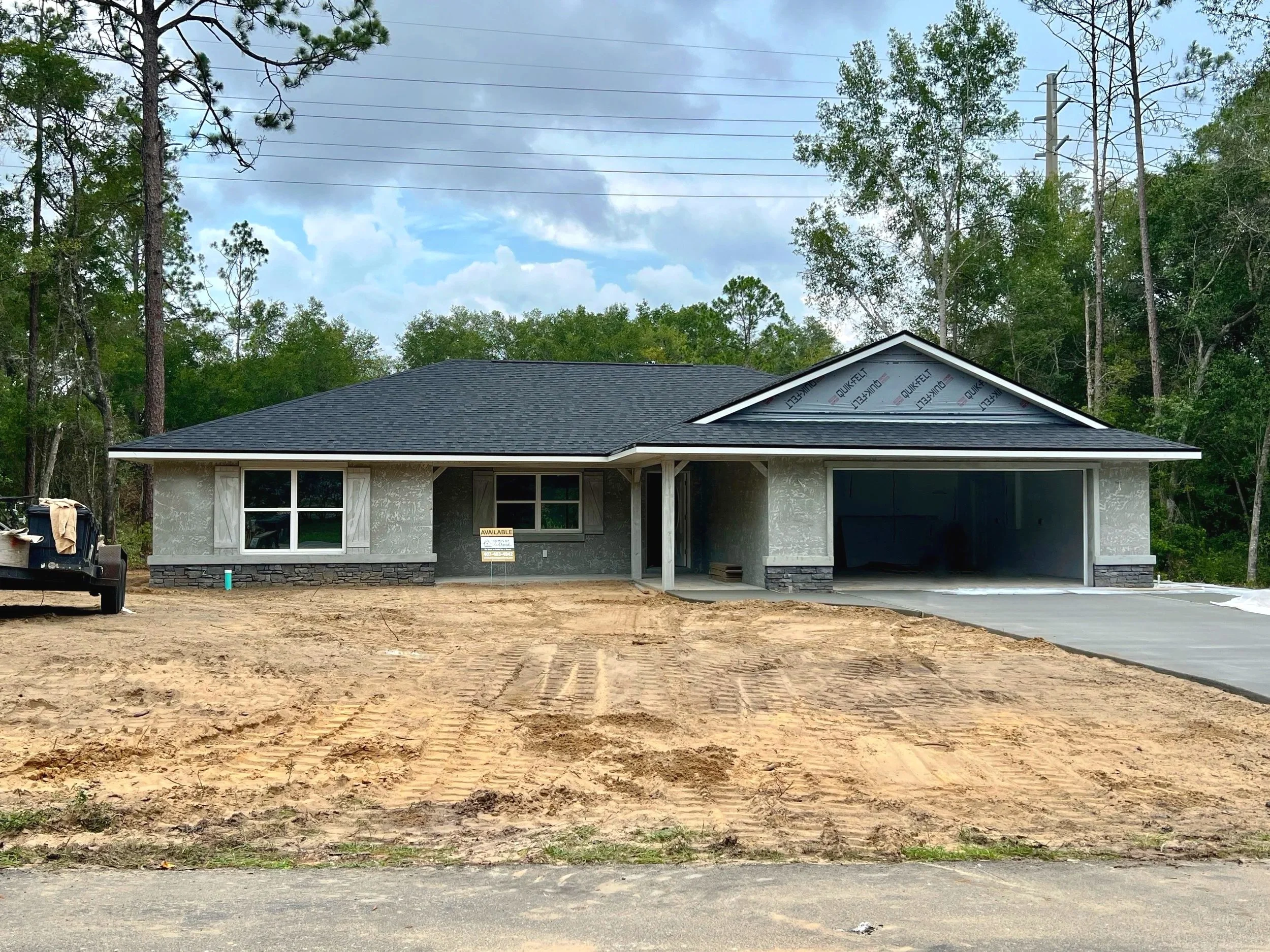 Newly constructed single-story gray stucco house under a cloudy sky, featuring dark roof shingles, stone accents along the base, an open two-car garage, and an unfinished dirt yard.