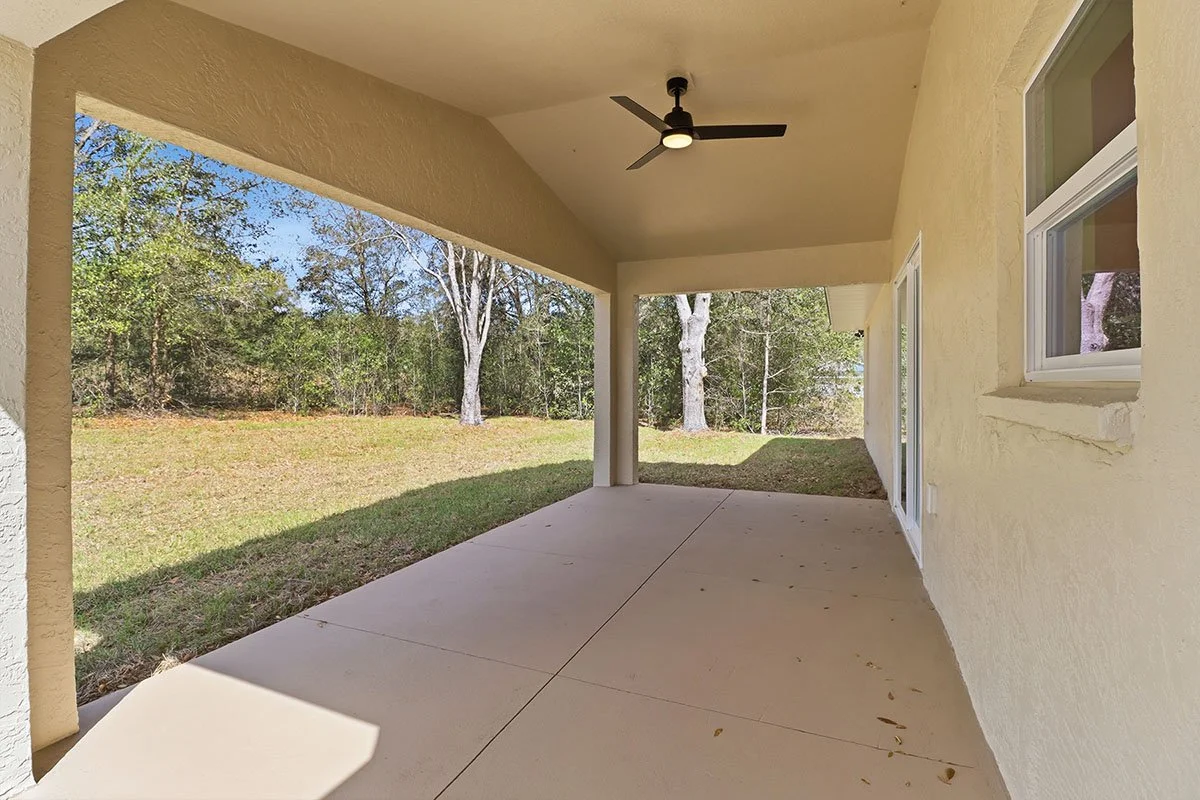 Covered backyard patio with ceiling fan, beige stucco walls, and a grassy yard with trees in the background.