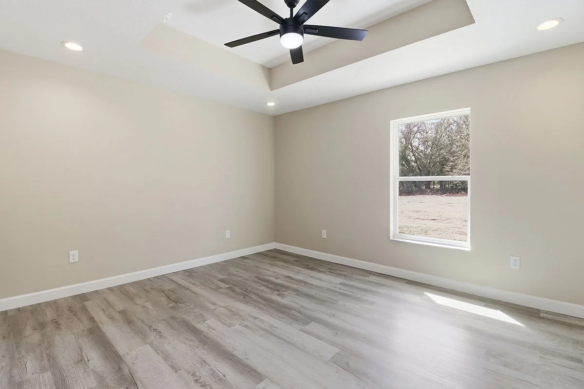Empty room with beige walls, a window showing an outdoor scene, a black ceiling fan with light, and wood-look flooring.