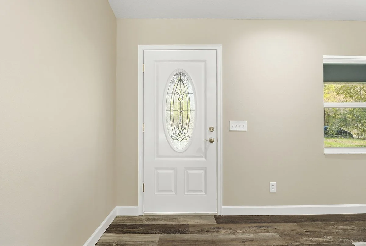 Interior view of a home entryway with a white door featuring an oval glass window with decorative design, beige walls, a window showing outdoor greenery, and wood-look flooring.