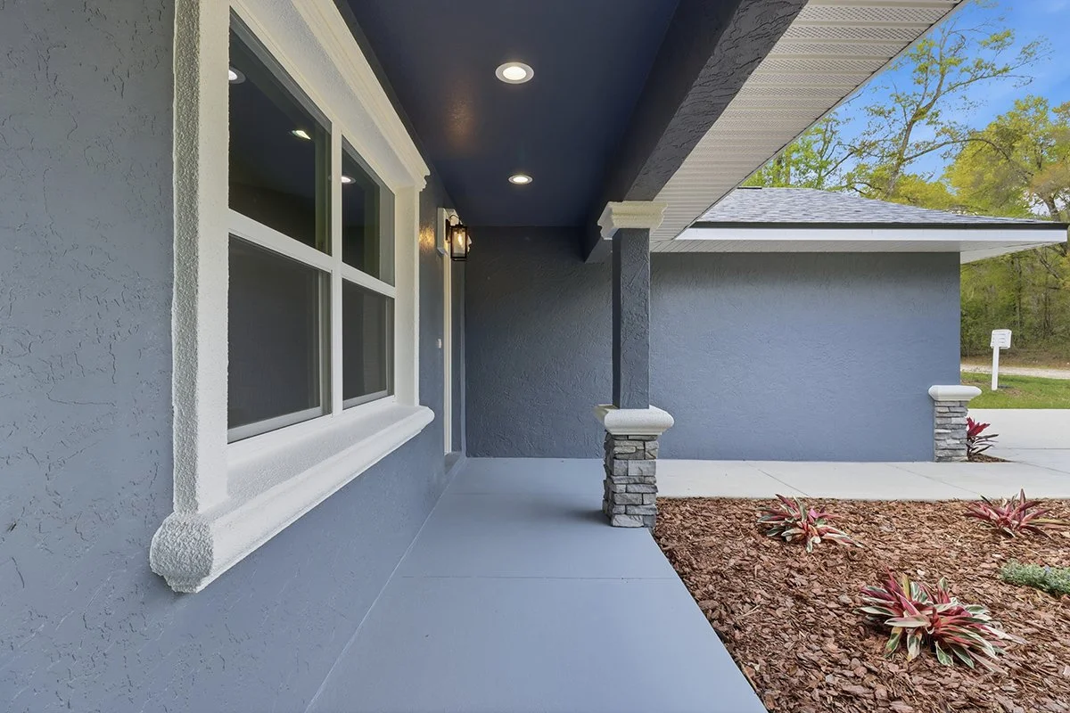 Front porch with gray textured walls, white window frame, small landscaped garden with reddish plants, and street in the background.