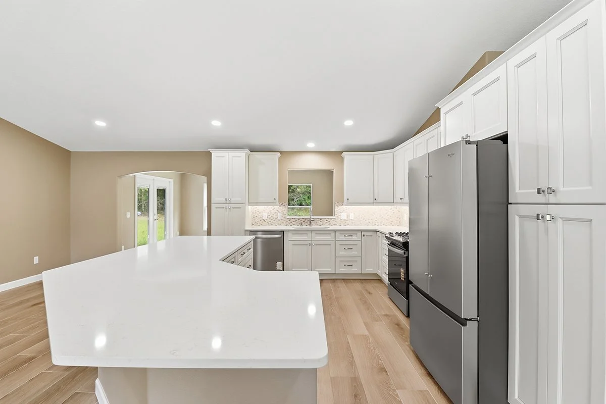 Modern kitchen with white cabinets, stainless steel refrigerator, black stove, and beige walls, featuring a large white island with a curved edge, wooden flooring, and recessed lighting.