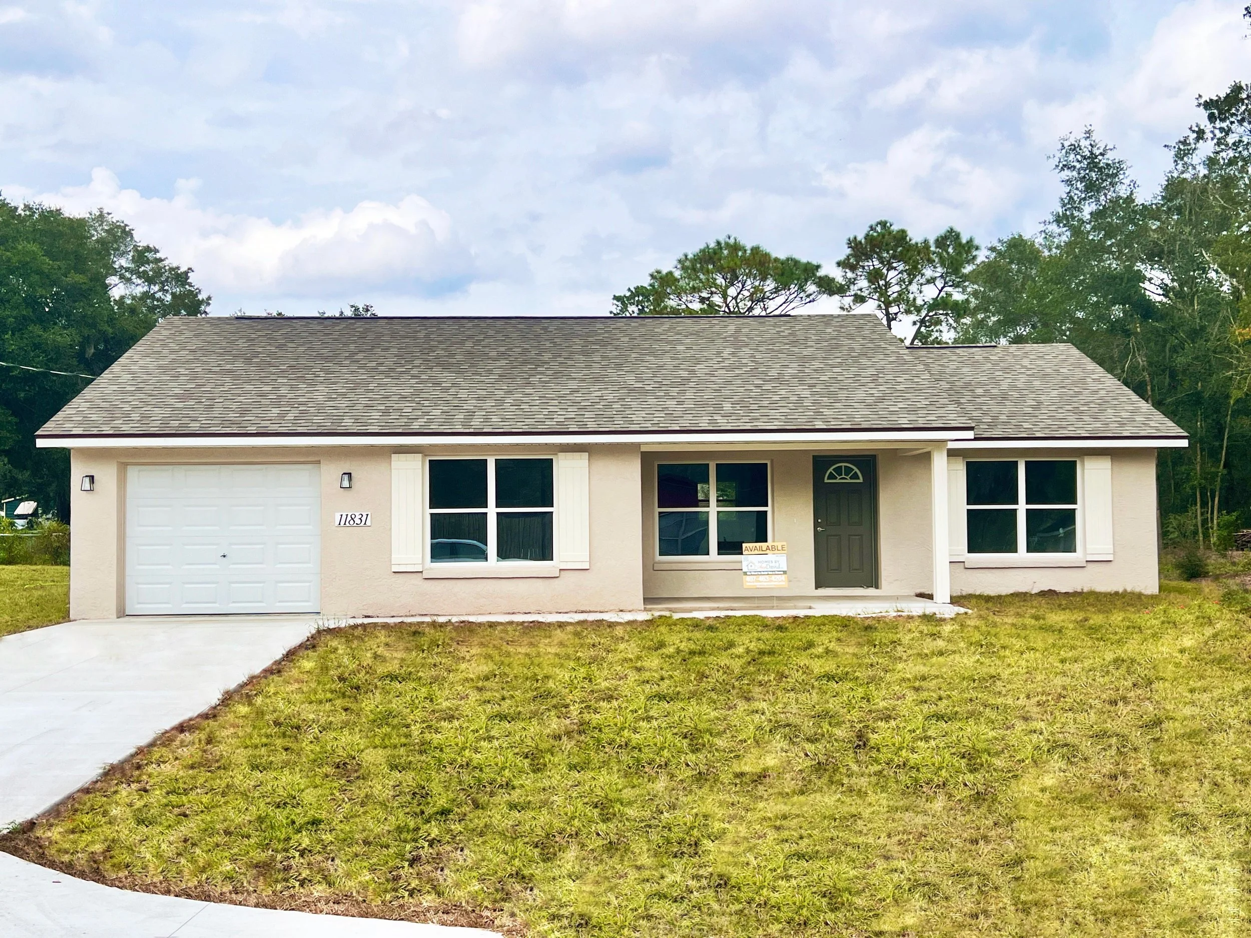Newly constructed home single family home with beige stucco exterior, beige shingles, a covered front porch with dark green front door, and a single car garage with white overhead door and concrete driveway.